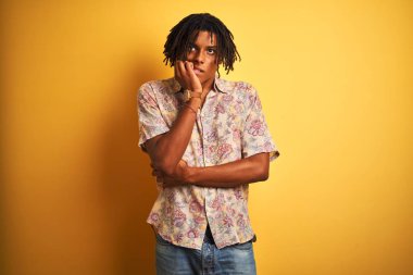 Tatilde dreadlocks ile Afro adam izole sarı arka plan üzerinde çiçek gömlek giyen stresli ve ağız tırnak ısırma elleri ile sinir görünümlü. Anksiyete sorunu.