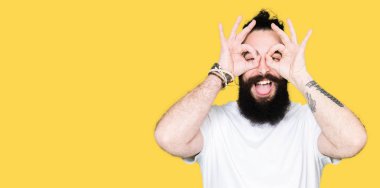 Uzun saç ve sakal, dilini parmaklarının arasından baktığını yapışmasını dürbün gibi ok jest yapıyor casual beyaz t-shirt giyen genç hipster adam. Deli ifade.