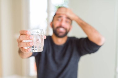 Yakışıklı İspanyol adam başında el ile vurguladı su taze bir bardak içme, utanç ve sürpriz yüz ile şok, kızgın ve sinirli. Korku ve hata için üzgün.