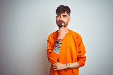 Dövmesi olan genç adam turuncu kazak giyen izole beyaz arka plan üzerinde duran çene üzerinde el soru, dalgın ifade düşünme. Düşünceli bir yüzle gülümsüyor. Şüphe kavramı.