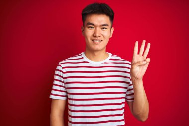 Genç asyalı Çinli adam çizgili t-shirt izole kırmızı arka plan üzerinde duran gösteren ve parmakları ile işaret ederken emin ve mutlu gülümseyerek üç.