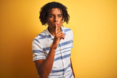 Izole sarı arka plan üzerinde çizgili gömlek giyen dreadlocks ile Afro Amerikan adam dudaklarında parmak ile sessiz olmak isteyen. Sessizlik ve gizli kavram.