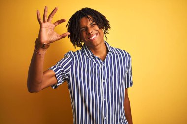 Izole sarı arka plan üzerinde rahat çizgili t-shirt giyen dreadlocks ile Afro adam gülümseyerek ve el parmakları görünümlü ve kamera ile küçük boyutlu işareti yapıyor kendinden emin gesturing. Ölçü kavramı.