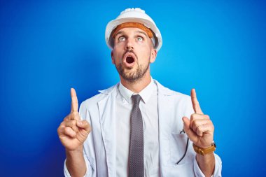 Mavi izole arka plan üzerinde güvenlik kaskgiyen genç yakışıklı mühendis adam şaşırttı ve baktı ve parmakları ile işaret ve kolları kaldırdı şaşırttı.