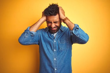 Denim gömlek giyen genç hintli adam izole sarı arka plan üzerinde ayakta baş ağrısı umutsuz muzdarip ve stresli çünkü ağrı ve migren. Eller baş üstünde.