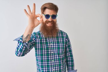 Genç kızıl İrlandalı adam izole beyaz arka plan üzerinde rahat gömlek ve güneş gözlüğü giyen olumlu el ve parmakları ile ok işareti yapıyor gülümseyerek. Başarılı ifade.