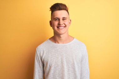 Genç yakışıklı adam yüzünde mutlu ve serin bir gülümseme ile izole sarı arka plan üzerinde duran beyaz rahat t-shirt giyiyor. Şanslı kişi.