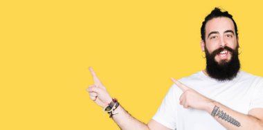 Uzun saçlı ve sakal ile genç hipster adam rahat beyaz t-shirt gülümseyen ve iki el ve parmak tarafına işaret kamera bakıyor.
