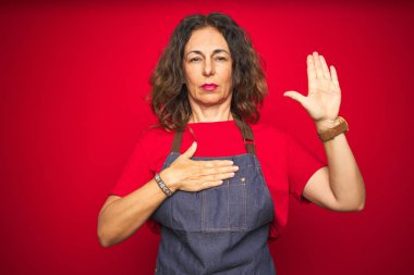 Orta yaş yaşlı kadın kırmızı izole arka plan üzerinde önlük üniforma giyen göğüs ve açık avuç içi el ile küfür, bir sadakat yemini yapma