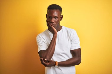 İzole sarı arka plan üzerinde duran beyaz t-shirt giyen genç afro-amerikan adam yorgun görünümlü ve çapraz kolları ile depresyon sorunları ile sıkılmış düşünme.