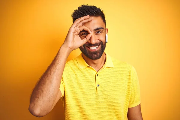 孤立した黄色の背景の上に立つイヤホンを使って音楽を聴いている若いインド人男性は 手を笑顔でOkジェスチャーを行い 幸せそうな顔で指を通して目を見る — ストック写真