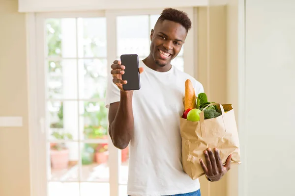 African man holding a paper bag full of groceries and using smarpthone buying online using app smiling