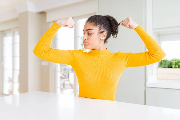 Beautiful african american woman with afro hair wearing a casual yellow sweater showing arms muscles smiling proud. Fitness concept.