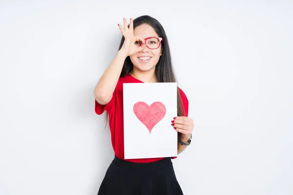 Young brunette woman holding card with red heart over isolated background with happy face smiling doing ok sign with hand on eye looking through fingers