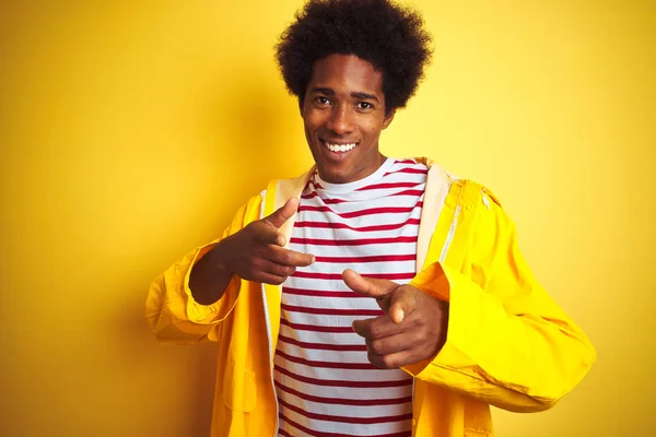 African american man with afro hair wearing rain coat standing over isolated yellow background pointing fingers to camera with happy and funny face. Good energy and vibes.