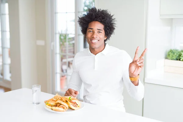 African American hungry man eating hamburger for lunch smiling with happy face winking at the camera doing victory sign. Number two.