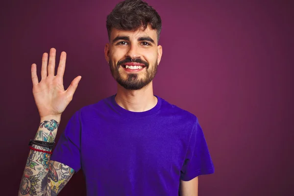 Young man with tattoo wearing t-shirt standing over isolated purple background showing and pointing up with fingers number five while smiling confident and happy.