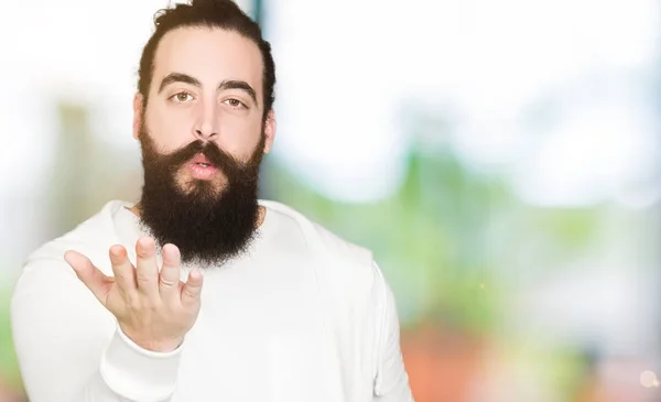 Young man with long hair and beard wearing sporty sweatshirt looking at the camera blowing a kiss with hand on air being lovely and sexy. Love expression.