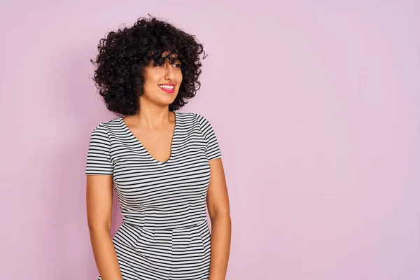 Young arab woman with curly hair wearing striped dress over isolated pink background looking away to side with smile on face, natural expression. Laughing confident.