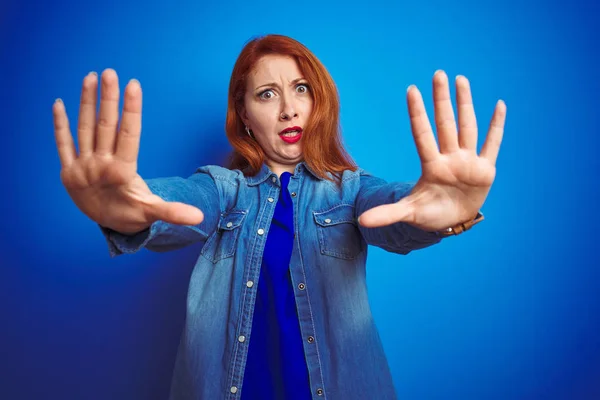 Young beautiful redhead woman wearing denim shirt standing over blue isolated background doing stop gesture with hands palms, angry and frustration expression