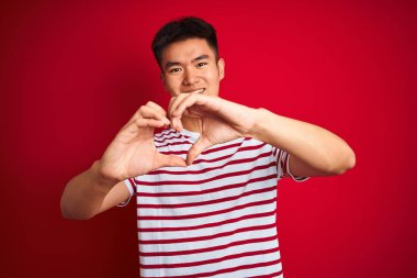 Genç asyalı Çinli adam çizgili t-shirt izole kırmızı arka plan üzerinde elleri ile kalp sembolü şekli yapıyor aşk gülümseyerek üzerinde duran giyiyor. Romantik kavram.