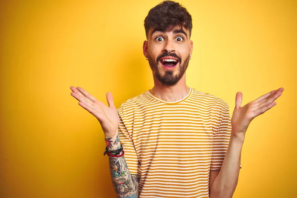 Çizgili t-shirt giyen dövme giyen genç adam izole sarı arka plan üzerinde duran deli kutluyor ve kollarını kaldırdı ve açık gözleri ile başarı için hayran heyecanlı çığlık. Kazanan kavramı