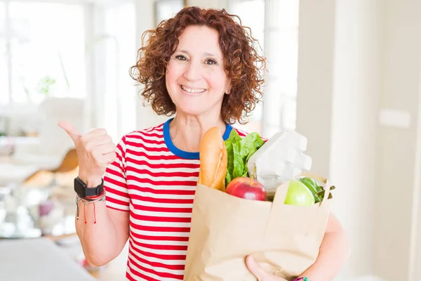 スーパーから新鮮な食料品がいっぱいの紙袋を持つ先輩女性が 笑顔で親指を立てて横に向かって見せる — ストック写真