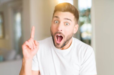 Evde rahat beyaz t-shirt giyen genç yakışıklı adam başarılı fikirle parmak işaret. Çıktıktan ve mutlu. Bir numara.