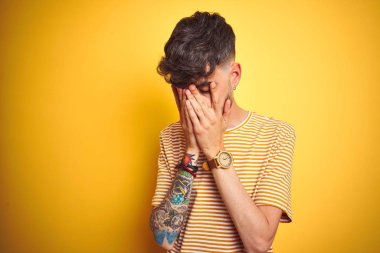 Dövmesi olan genç adam, izole edilmiş sarı arka plan üzerinde duran çizgili tişört giyerken, üzgün ifadesi ile elleri ile yüzü kaplayarak ağlarken. Depresyon kavramı.