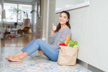 Genç kadın bir kağıt çanta taze bakkaliye dolu ile mutfak katında oturan mutlu thumbs el ile jest yapıyor. Başarı göstererek kameraya bakarak ifadeyi onaylama.