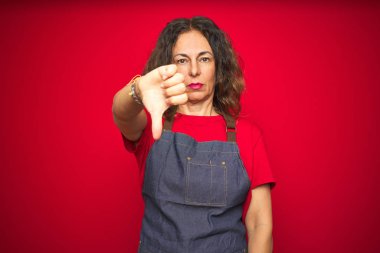 Kırmızı izole arka plan üzerinde önlük üniforma giyen orta yaş yaşlı kadın mutsuz ve kızgın görünümlü reddedilme ve aşağı jest başparmak ile olumsuz gösteren. Kötü ifade.