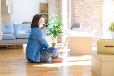 Genç artı boyutu kadın karton kutular etrafında yan bakarak yeni bir eve hareket etrafında katta oturan, kendine güvenen gülümseme ile doğal yüz ile profil poz rahatla.