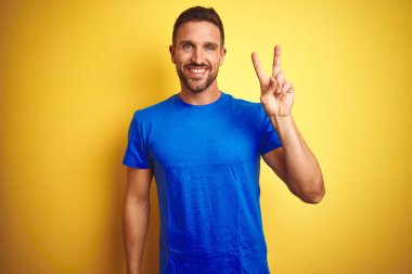 Sarı izole edilmiş arka plan üzerine sıradan mavi tişört giyen genç yakışıklı adam kameraya gülümsüyor ve zafer işareti yapıyor. İki numara..