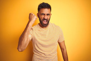 İzole sarı arka plan üzerinde duran t-shirt giyen genç hintli adam kızgın ve öfkeli yükselterek yumruk sinirli ve öfkeli öfke ile bağırırken. Öfke ve agresif kavram.