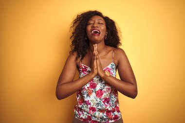 Izole sarı arka plan üzerinde çiçek yazlık t-shirt giyen Afrikalı-Amerikalı kadın dilencilik ve yüz umut ifadesi ile birlikte elleri ile dua çok duygusal ve endişeli. Af dilemek istiyorum. Din kavramı.