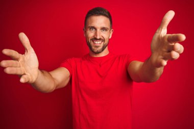 Genç yakışıklı adam kırmızı izole arka plan üzerinde rahat t-shirt giyen sarılmak için açık kollarla gülümseyerek kameraya bakarak. Mutluluğu kucaklayan neşeli ifade.