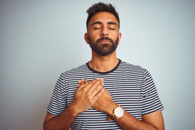 Siyah çizgili t-shirt giyen genç hintli adam kapalı gözleri ve yüzünde minnettar jest göğüs elleri ile gülümseyerek izole beyaz arka plan üzerinde duran. Sağlık kavramı.