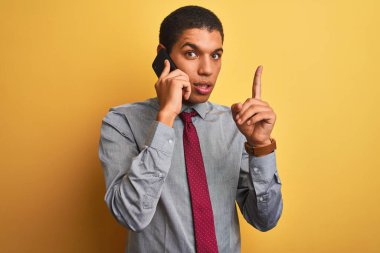 İzole sarı arka plan üzerinde akıllı telefonda konuşan genç yakışıklı Arap işadamı mutlu yüz, bir numara ile bir fikir ya da soru işaret parmağı ile şaşırttı