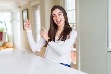 Güzel genç kadın evde beyaz masada gülümseyerek oturan ve yan iki el ve parmakları ile işaret kameraya bakıyor.