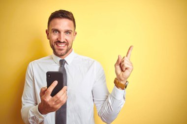 Genç yakışıklı iş adamı sarı izole arka plan üzerinde akıllı telefon kullanarak çok mutlu yan el ve parmak ile işaret