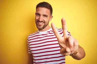 Genç yakışıklı adam sarı izole arka plan üzerinde rahat kırmızı çizgili t-shirt giyen kameraya bakarak gülümseyerek zafer işareti yapıyor parmakları gösteren. İki numara.