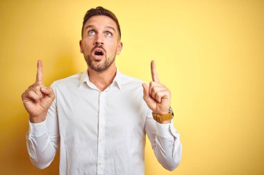 Genç yakışıklı adam sarı izole arka plan üzerinde zarif beyaz gömlek giyen şaşırmış ve şaşırmış yukarı bakarak ve parmakları ile işaret ve kolları kaldırdı.