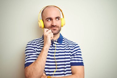 Young man listening to music wearing yellow headphones over isolated background with hand on chin thinking about question, pensive expression. Smiling with thoughtful face. Doubt concept. clipart