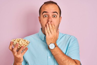 Antep fıstığı ile kase tutan genç adam el ile izole pembe arka plan kapağı ağız üzerinde duran hata için utanç ile şok, korku ifadesi, sessizlik, gizli kavram korkmuş
