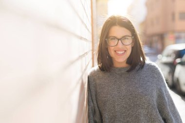 Gözlük giyen güzel genç kadın neşeli tuğla duvara eğilerek gülümseyen, şehirde rahat güzel kız