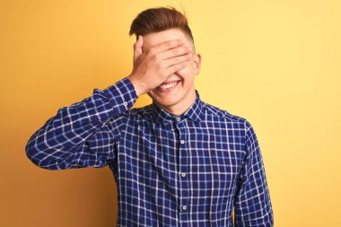 Sarı arka plan üzerinde dikilen rahat tişörtlü genç yakışıklı adam. Gülümsüyor ve elleri yüzünde şaşkınlıktan gözleri kapatıyordu. Kör kavram.