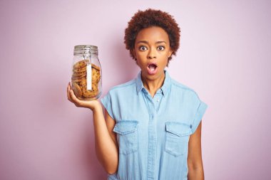 Pembe izole arka plan üzerinde çikolata cipsi kurabiye kavanoz tutan Afrikalı Amerikalı kadın sürpriz bir yüz ile şok korkmuş, korku ifade korku ve heyecanlı
