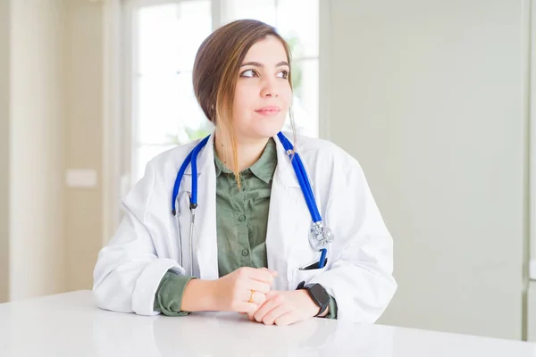 Beautiful young doctor woman wearing medical coat and stethoscope looking to side, relax profile pose with natural face with confident smile.