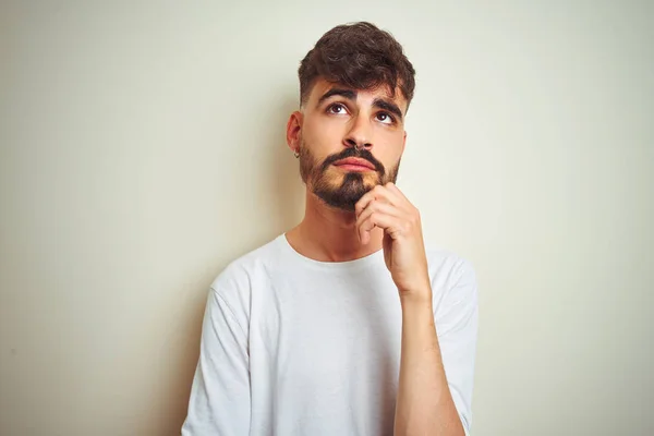 Young man with tattoo wearing t-shirt standing over isolated white background with hand on chin thinking about question, pensive expression. Smiling with thoughtful face. Doubt concept.