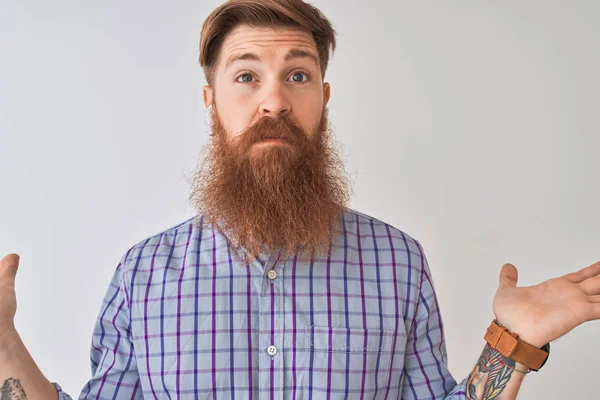 Redhead irish man listening to music using wireless earphones over isolated white background clueless and confused expression with arms and hands raised. Doubt concept.